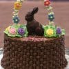 The EA-025 is a chocolate cake featuring basket-weave frosting, a chocolate bunny, colorful icing flowers, green leaves, and an arch crafted from decorative icing flowers.