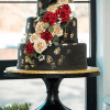 The WD-000C is a three-tiered black cake with gold accents, cascading red and cream roses, greenery, and small white flowers, displayed on a black cake stand by a window.