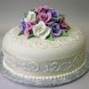 AN-027 is a round white cake topped with lifelike purple, pink, and white fondant roses, green leaves, and elegant white icing swirls, presented on a silver cake board.