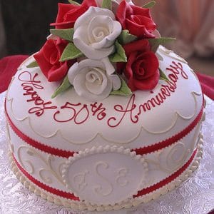 The AN-017 is a round white cake adorned with red and white roses, gold scalloped icing, and a red ribbon. "Happy 50th Anniversary" is written in red script on top, with the initials S E on the side.