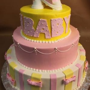 The BB-001 is a two-tiered baby shower cake in pink, yellow, and white, topped with fondant baby shoes and large BABY letters. The lower tier features pink and yellow bows with stripe accents.