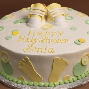 BB-021 is a round baby shower cake with yellow and green decorations, baby shoe toppers, footprint shapes on the sides, and "Happy Baby Shower Sonia" written in yellow icing on top.