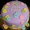 BB-066 is a round cake with purple fondant, decorated with pastel pink, yellow, green, and blue fondant buttons, and "Cute as a Button" written in white icing on top.