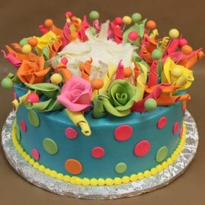 The BD-057 cake features blue frosting, vibrant pink, orange, green, and yellow fondant flowers and dots, finished with a yellow icing border at the base and presented on a foil-covered board.