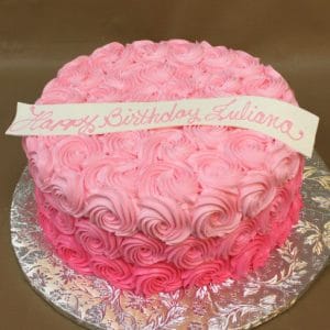 CB-018D is a round cake adorned with pink rosette frosting and topped with a white banner reading "Happy Birthday Juliana" in pink script, displayed on a silver floral-patterned cake board.