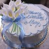 BR-038 is a round white cake with blue icing, a blue ribbon, and white calla lilies. "Happy Birthday Mary" is written in blue script on top. The cake is presented on a gold board.