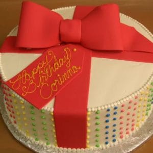 CB-023 is a round birthday cake with white frosting, colorful dot decorations on the sides, a large red fondant bow, and a yellow tag that says "Happy Birthday Corinna.