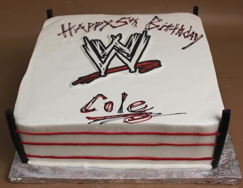 The ST-010 Wrestling cake is rectangular and decorated as a wrestling ring with red ropes, black corner posts, the WWE logo, and "Happy 5th Birthday Cole" written in black and red icing.