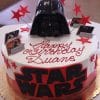 Product CB-007A is a round Star Wars-themed birthday cake with large black and red “STAR WARS” lettering, a Darth Vader helmet topper, red stars, small Star Wars cards, and “Happy Birthday Duane” in red icing.