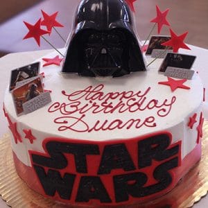 Product CB-007A is a round Star Wars-themed birthday cake with large black and red “STAR WARS” lettering, a Darth Vader helmet topper, red stars, small Star Wars cards, and “Happy Birthday Duane” in red icing.