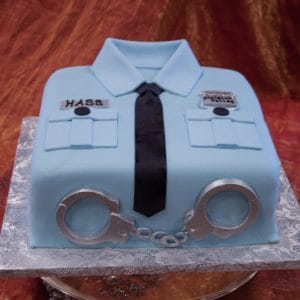 GC-053 is a square cake designed as a light blue police uniform shirt with a black tie, silver handcuffs, name badge, and pocket details, set on a silver-patterned cake board.