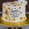 BR-000J is a white cake with gold polka dots, topped with a large white flower and a heart-shaped "Cheers to Love" plaque. It features a gold-edged champagne glass design on the side and is presented on a gold cake stand.