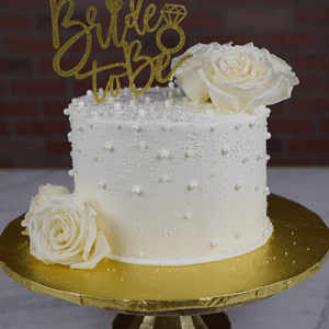 The BR-000I is a white cake adorned with pearls and white roses, featuring a gold "Bride to Be" sign and diamond ring design, displayed on a gold stand against a brick wall background.