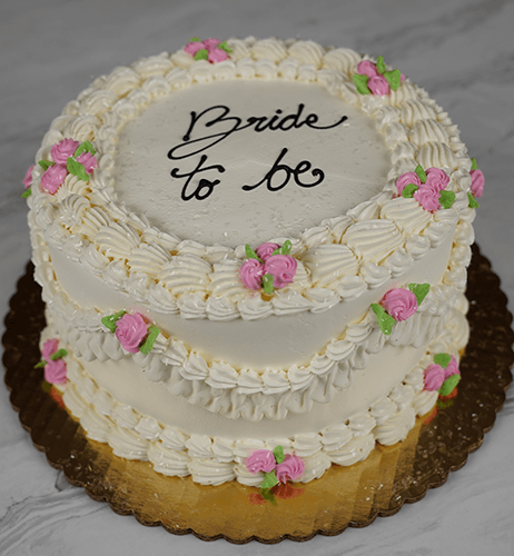 The BR-000H is a two-tiered white frosted cake with pink and green icing flowers, piped borders, and "Bride to be" written in black icing on top. It sits on a gold board.