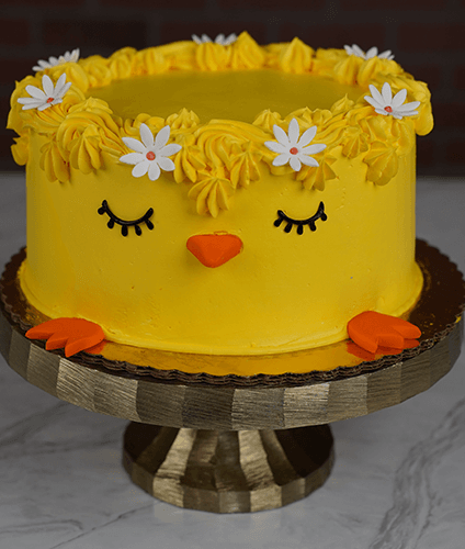 EA-000C is a round yellow cake designed like a chick, featuring closed eyes, an orange beak and feet, with a crown of yellow frosting and white flowers on top, displayed on a cake stand.