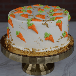 The EA-000D Carrot cake is a round cake topped with white frosting, orange and green icing carrots, and sprinkled crumbs on top and around the base, presented on a gold cake stand.