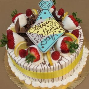 The FD-009 Gold cake features a round shape with chocolate shavings, lemon slices, and strawberries—some dipped in white chocolate. A blue tie-shaped plaque says "Happy Fathers Day." The gold cake has a lemon and raspberry mousse filling and sits on a gold board.