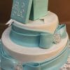 The TC-105 is a two-tier white cake with blue fondant ribbon details, jeweled brooches, and a monogrammed blue gift box with a white bow on top, displayed on a silver-patterned board.