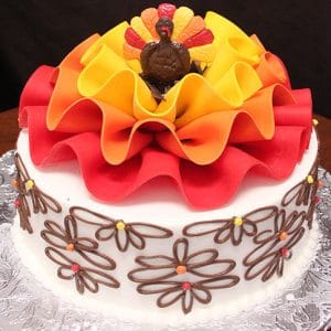 The THX-026 cake is round with white icing, decorated with chocolate flower piping, colorful dots, large red, orange, and yellow ribbon-like fondant on top, and a small turkey figure at the center.