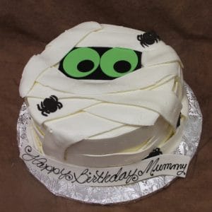 The HW-007 is a round cake designed as a mummy with white icing "bandages," green and black eyes, tiny black spiders, and "Happy Birthday Mummy" written at the base.