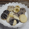 A white scalloped platter showcases CO-045 Assorted Tea Cookies—including chocolate-dipped, powdered sugar, nut-topped, and jam-filled varieties—elegantly arranged on a rustic wooden surface.