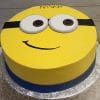 The CB-013B is a round cake styled as a Minion with yellow frosting, big white and brown eyes, a black goggles strap, smiling mouth, and a blue band at the base.