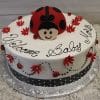 CB-012I: Round cake with a large fondant ladybug, red flowers, small ladybugs, and “Welcome Baby Mia” in black icing on white frosting, finished with a black polka dot ribbon around the base.