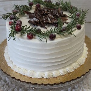 The CH-029 is a white frosted cake topped with a rosemary wreath, sugared cranberries, and chocolate shavings, presented on a gold cake board with draped white fabric.