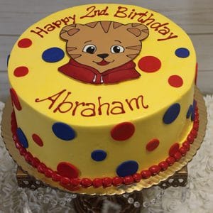 The CB-009O is a yellow cake with red and blue polka dots, a cartoon tiger face, and red piping. "Happy 2nd Birthday" is written on top and "Abraham" appears in red icing on the side.