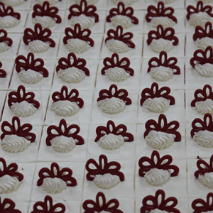 Neatly arranged CO-003 Red Velvet 1X1 Bites feature square pastries topped with white cream swirls and dark red decorative icing, all set on a white frosting background.