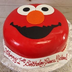 The CB-003C is a round cake designed as Elmo’s face with red icing, white eyes, an orange nose, and a black mouth. The cake board features the message: “Happy 1st Birthday Prince Michael!”.