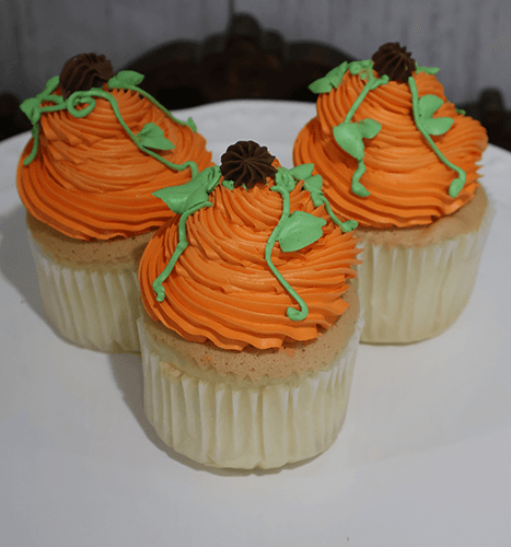 Three CC-072 Pumpkin design cupcakes, featuring gold cake with chocolate mousse filling, are decorated as pumpkins with orange frosting, green vines, and brown stems, and arranged on a white plate.