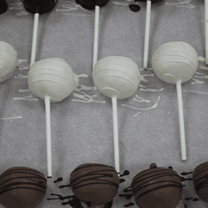 CO-032 Chocolate Cake Pops—rows of cake pops on sticks, coated in dark, white, and milk chocolate with decorative chocolate drizzle, arranged on parchment.