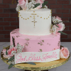 The RC-001B is a two-tiered cake with pink and white frosting, pink roses, white flowers, gold cross and accents. A banner reads “God Bless Janelle.” Presented on a gold stand.