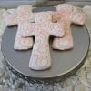 Three CE-005a religious cross-shaped cookies with white icing and pink swirls sit on a round silver stand with a lacy edge, displayed on ruffled white fabric.
