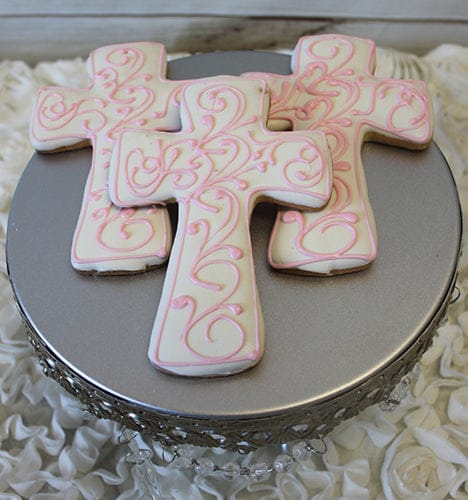 Three CE-005a religious cross-shaped cookies with white icing and pink swirls sit on a round silver stand with a lacy edge, displayed on ruffled white fabric.