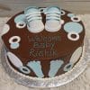 BB-023 is a round chocolate cake adorned with blue and white fondant baby shoes, circles, and footprints, finished with "Welcome Baby Rishik" written in blue icing.