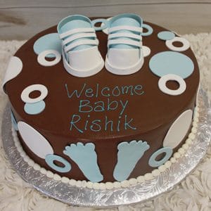 BB-023 is a round chocolate cake adorned with blue and white fondant baby shoes, circles, and footprints, finished with "Welcome Baby Rishik" written in blue icing.