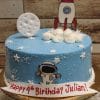 CB-027C is a blue birthday cake with white stars, a moon, an astronaut, and a cartoon rocket. It features a sign in red letters that reads, "Happy 4th Birthday Julian!.