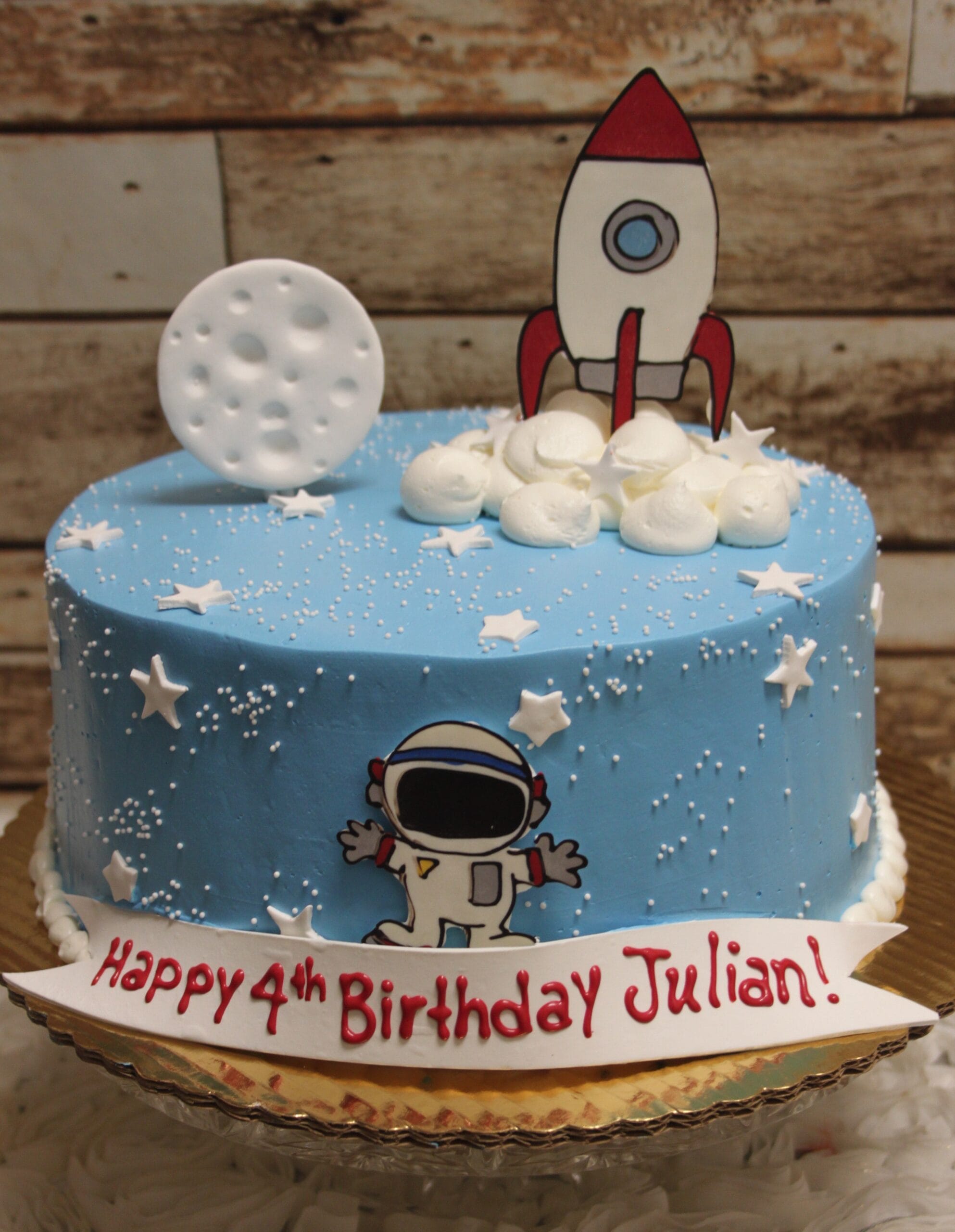 CB-027C is a blue birthday cake with white stars, a moon, an astronaut, and a cartoon rocket. It features a sign in red letters that reads, "Happy 4th Birthday Julian!.