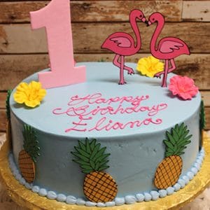 The CB-020B is a round blue birthday cake topped with yellow and pink flowers, pineapples, two pink flamingos, a large pink number 1, and “Happy Birthday Eliana” written in pink icing.