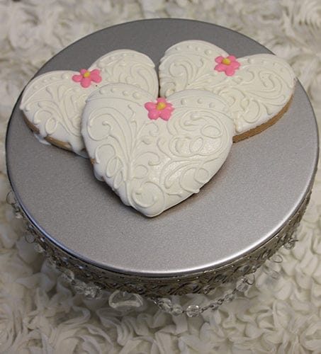 Three CE-003c bridal heart-shaped cookies with white ornate icing and pink flowers are displayed on a silver cake stand with decorative trim, set against a textured white background.