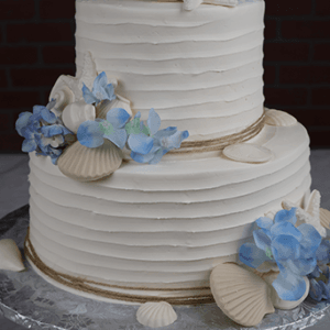 The WD-001R is a two-tiered white cake adorned with blue hydrangea flowers, white seashells, and textured frosting. It’s presented on a silver cake board with a gold stand, set against a dark brick background.