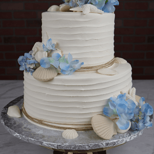 BR-000E: Two-tiered white cake adorned with blue hydrangea flowers, white seashells, and twine accents, presented on a silver cake board and brass stand against a dark brick background.