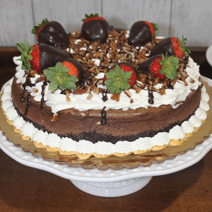 GD-007 Turtle cheesecake topped with whipped cream, chopped nuts, chocolate drizzle, and chocolate-covered strawberries, displayed on a white cake stand.
