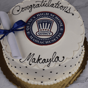A round white cake (GC-001C Quincy High School) features "Congratulations Makayla" in black icing, a Quincy High emblem at the center, and a rolled diploma with a blue ribbon on top. Displayed on a gold board.