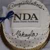 A round GC-001F Notre Dame Academy cake with "Congratulations Makayla" in icing, the NDA logo, and a small rolled diploma tied with a blue ribbon on top.