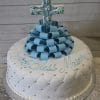 The RC-076 is a round white cake with quilted fondant, silver pearls, blue fondant loops, and a blue cross on top. “God Bless Mason” is written in blue icing.