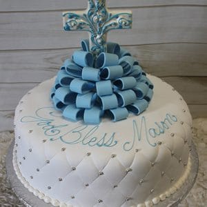 The RC-076 is a round white cake with quilted fondant, silver pearls, blue fondant loops, and a blue cross on top. “God Bless Mason” is written in blue icing.