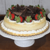 The GD-001 New York Style Cheesecake, topped with chocolate shavings and chocolate-dipped strawberries and decorated with whipped cream, is displayed on a scalloped-edge white cake stand.